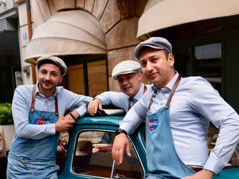 Genusshelden: drei Metzger in Schürzen und Schiebermützen an einem türkisen Oldtimer.