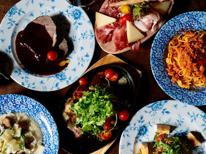 Genusstour: gedeckter Tisch von oben, Braten, Pasta, Antipasti und Risotto auf blau gemusterten Tellern.