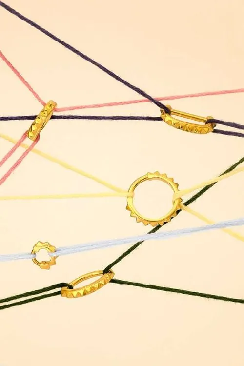 Goldene Piercing-Ringe von Giada Ilardo, aufgefädelt auf farbigen Fäden vor pastellfarbenem Hintergrund.